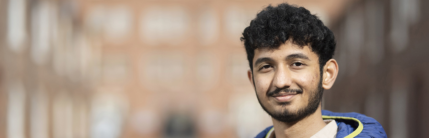 A young man wearing a blue puffer jacket and a light-colored shirt, stands outdoors in a blurred urban setting, smiling at the camera.