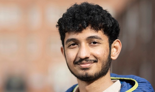 A young man wearing a blue puffer jacket and a light-colored shirt, stands outdoors in a blurred urban setting, smiling at the camera.