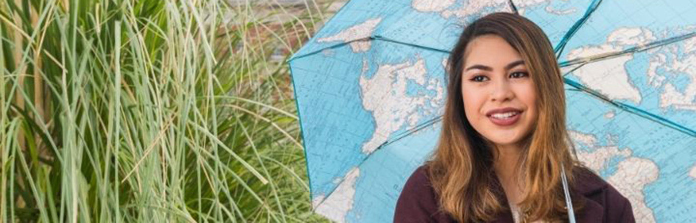 A young woman with shoulder-length brown hair smiling and holding a blue umbrella featuring a world map design, standing in front of tall green grass.