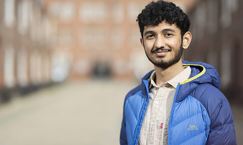 A young man wearing a blue puffer jacket and a light-colored shirt, stands outdoors in a blurred urban setting, smiling at the camera.