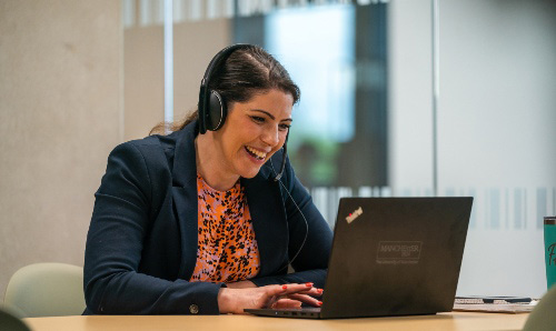 A woman wearing headphones and a blazer smiles while engaged in a video call on her laptop.