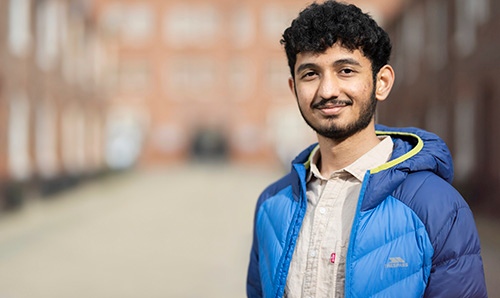 A young man wearing a blue puffer jacket and a light-coloured shirt, stands outdoors in a blurred urban setting, smiling at the camera.